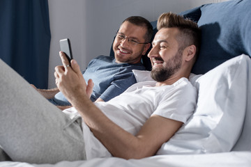 Happy gay couple using smartphone while lying together on bed