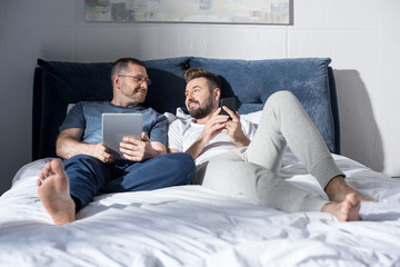 Happy boyfriends using smartphone and digital tablet while lying together in bed and smiling each...