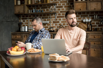 Serious gay couple using laptop and smartphone while sitting at table with fresh breakfast