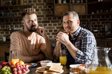 Happy gay couple sitting at table with tasty breakfast and smiling at camera