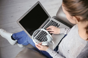 Woman using laptop with headphones while sitting in arm-chair. Concept of audiobook