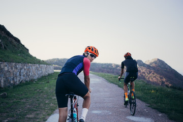 Two friends enjoy riding bikes together on empty mountain summit in soft summer sunset light, one cyclist looks back on camera happy and amazed by view
