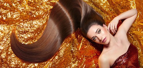 Naklejka premium A beautiful young girl with long shiny silky hair lies in a shiny dress on a gold background. Cosmetics for hair, care, spa, beauty, fashion. Beauty saloon. Smooth skin. Advertising.Shine, radiance.