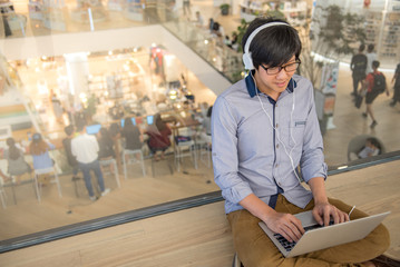 Young Asian man dressed in casual style using laptop computer while listening to music. Digital nomad working in co working space, modern IT lifestyle with work life balance concept.