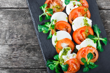 Caprese salad with Mozzarella cheese, tomatoes, pesto sauce and basil herb leaves on black slate stone chalkboard on wooden background close up. Mediterranean healthy food. Top view