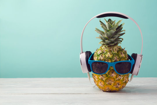 Pineapple with headphones and sunglasses on wooden table over mint background. Tropical summer vacation and beach party concept.