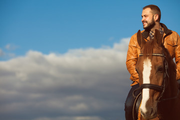 Country life concept. Young relaxed and smiling rich man in light brown leather jacket riding a horse. Sunny cloudy weather. Wide open spaces. Copy-space. Retro style. Outdoor shot