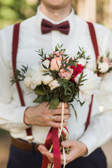 Closeup of young handsome stylish groom holding perfect bridal bouquet of fresh flowers in hands....