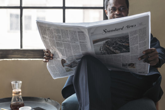Middle-aged African businessman sitting on armchair and reading newspaper.