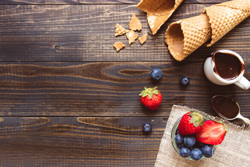 fresh berries, waffle cone and chocolate sauce on the wooden background, top view. Ice-cream background with copy space