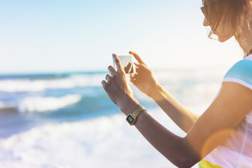 Hipster girl photograph on smart phone gadget in sand coastline, mock up of blank screen. Traveler hold and using in female hand mobile on background seascape horizon blue sun ocean, lifestyle