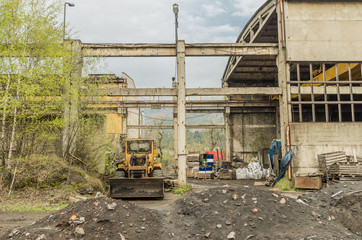 Fototapeta na wymiar Outside view of old abandoned factory ruin with broken windows and glass rusty steel structure with yellow bulldozer.