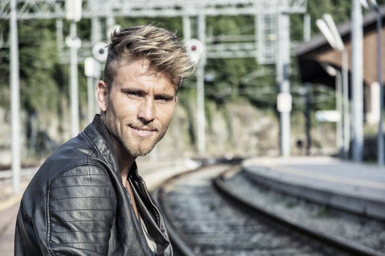 Attractive blond young man standing on railroad tracks, wearing only black leather jacket, looking at camera