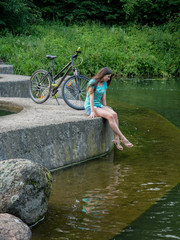 A beautiful girl is sitting near a bicycle near a pond.