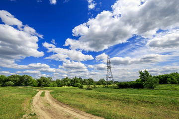 road and green field with trees and blue sky with clouds sunny day, beautiful rural landscape. Electricity tower standard overhead power line transmission tower