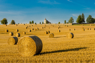 Romantic view of straw bales field. Sunlit summer landscape after a harvest of grain with a church and a churchyard on the horizon. Scenic rural background of golden stubble, blue sky and green trees.