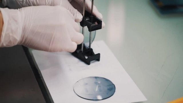 Scientist in white rubber gloves puts wet glass lenses on paper sheet upon glossy desk in laboratory, close up