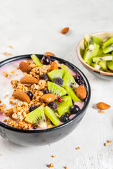 Healthy diet breakfast: smoothies bowl, with yogurt, fresh blueberries (black currant), kiwi, granola oatmeal, almond nuts. On a white stone table, with a glass of water and ingredients. Copy space