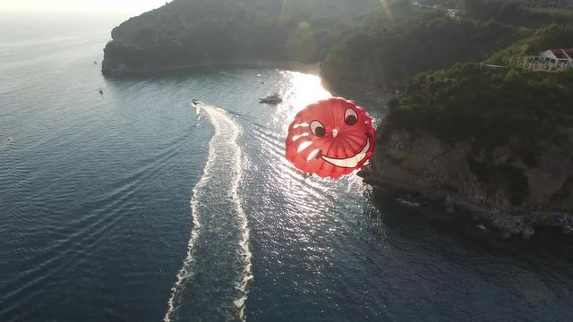 Red smile parachute. Paragliding behind a motor boat In Montenegro bay Budva. Extreme sport on vacation trip. Aerial drone follow people on the rope. Close flight. Summer sunset