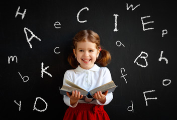 Happy schoolgirl preschool girl with book near school blackboard