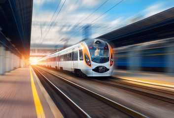 Obraz premium Szybki pociąg w ruchu na stacji kolejowej o zachodzie słońca w Europie. Nowoczesny pociąg międzymiastowy na peronie kolejowym z efektem rozmycia ruchu. Scena przemysłowa z poruszającym się pociągiem pasażerskim na kolei