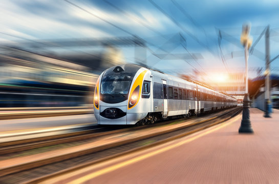 Fototapeta High speed train in motion at the railway station at sunset. Modern european intercity train on the railway platform with motion blur effect. Industrial scene with moving passenger train on railroad