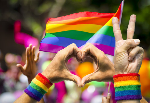 Supporting hands make peace and heart signs in front of a rainbow flag flying on the sidelines of a summer gay pride parade