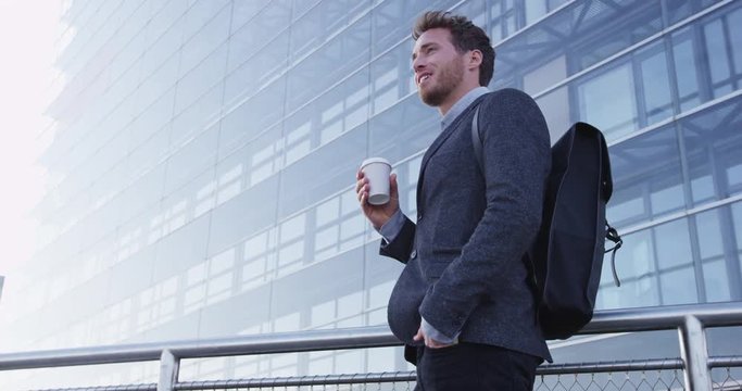 Businessman portrait of young male urban professional business man in suit standing outside office building. Confident successful young caucasian man drinking coffee wearing cool backpack. SLOW MOTION