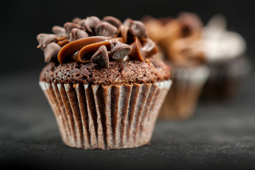 Three chocolate cupcakes garnished with chocolate chips and frosting on lined up across the frame, on black background