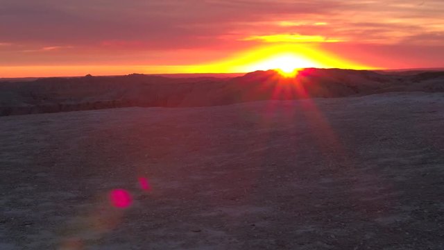 AERIAL: Beautiful reddish sun setting behind the endless sandstone mountains at Badlands National Park at sunset. Stunning sunbeams shinning over eroded rocky mountaintop formations at sunrise in USA