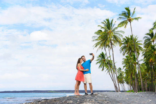 Selfie couple on Hawaii beach vacation with palm trees and volcanic black sand in Big island of Hawaii, USA. Hawaiian holidays getaway. Happy people on summer holidays.