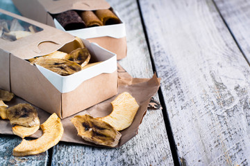 Paper box with fruit apple and banana chips on wooden table background. Sweet vegan healthy dessert. Copy space, top view
