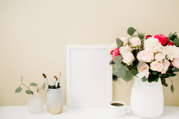 Home office desk with photo frame mockup, beautiful roses and eucalyptus bouquet in front of pale pastel beige background. Blog, website or social media concept .