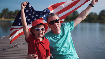Father with son holding American flag and sitting on a pier near water celebrating 4th July or Fathers day