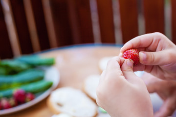 Close-up photo of boy´s hands as he torns a stem from strawberry, before eating it. Photo is taken on terace, next to a table.