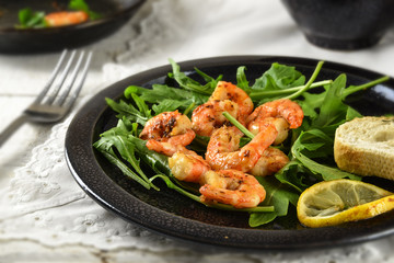 roasted prawns or shrimps on arugula rocket salad with lemon and bread served on a black plate, white tablecloth