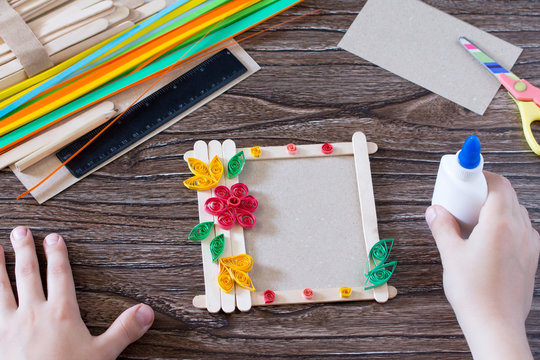 The child glues the details to a gift photo frame made of wooden sticks. Handmade. Project of children's creativity, handicrafts, crafts for children.