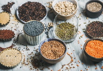 Various raw uncooked grains, beans, cereals in bowls, cups and glass for healthy cooking over marble background, selective focus. Clean eating, dieting, healthy, detox, vegan, vegetarian food concept