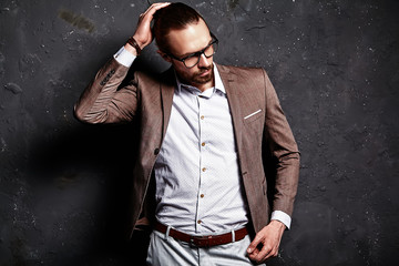 portrait of handsome fashion stylish hipster businessman model dressed in elegant brown suit in glasses posing near dark wall in studio