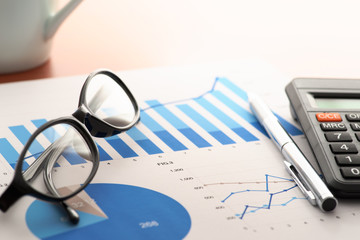 Checking report on business table. Glasses, pen, calculator and document with blue charts on wooden desk.
