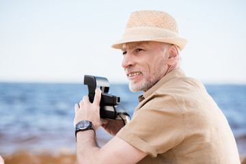 Obraz premium Smiling senior man in hat holding instant camera and looking away outdoors