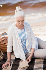 Fototapeta na wymiar Pensive senior woman sitting on plaid with book and picnic basket