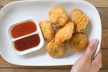 hand of young woman holding dish with chicken nuggets. have small cup with chilli sauce and tomato sauce all of it putting on wooden table are background. this image for food concept