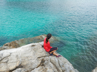 asia woman wearing red shirt sitting on rock and looking scenery view. front of her have blue sea and green mountain are background. this image for nature,seascape,travel and portrait concept