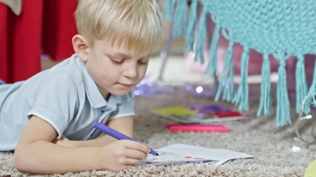 Closeup of cute little boy lying on carpet on the floor and drawing handmade greeting card