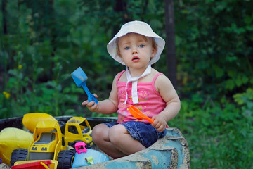 A small charming girl is playing in the sandbox in the backyard of her house. Infant is sitting with a plastic spatula among toys in a sandbox made of a large wheel from a tractor.