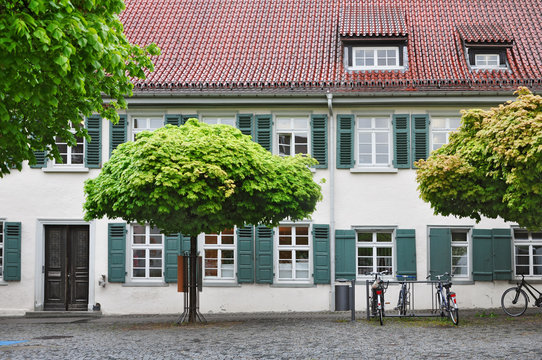 An old building with wooden windows and shutters. Decorative green maples and parked bicycles in the foreground.