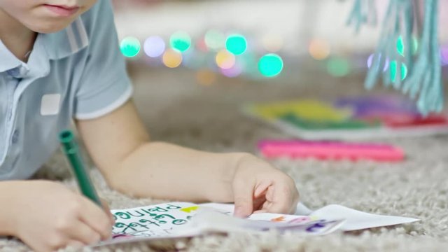 Tracking shot of little boy lying on the floor and writing wishes in greeting card with felt tip pen
