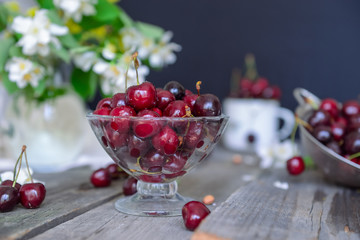 Fresh cherry fruit in glass vase, other dishes with berries and jar with jasmine and wildflowers on the old wooden table. Soft selective focus. Summer rustic concept