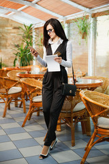 A business woman is standing in a cafe with a mobile phone and a notebook in her hands